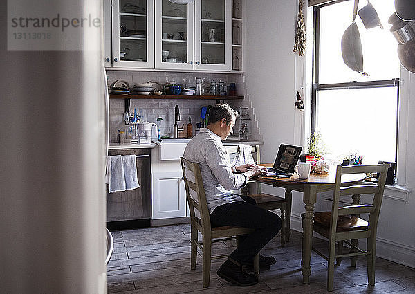 Seitenansicht eines Mannes  der einen Laptop-Computer benutzt  während er am Esstisch in der Küche sitzt