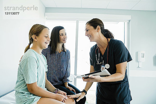 Fröhliche Kinderärztin im Gespräch mit einem Mädchen  das neben der Mutter auf dem Untersuchungstisch im Krankenhaus sitzt