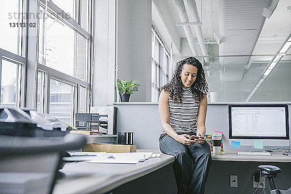 Geschäftsfrau benutzt Mobiltelefon  während sie im Büro am Schreibtisch sitzt