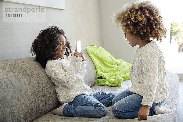 Fröhliches Mädchen fotografiert Schwester mit Smartphone auf Sofa