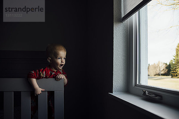 Kleiner Junge mit offenem Mund  der durch ein Fenster schaut  während er zu Hause am Stuhl steht