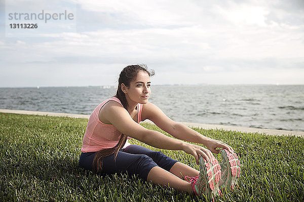 Junge Frau berührt Zehen  während sie auf Grasfeld gegen das Meer sitzt