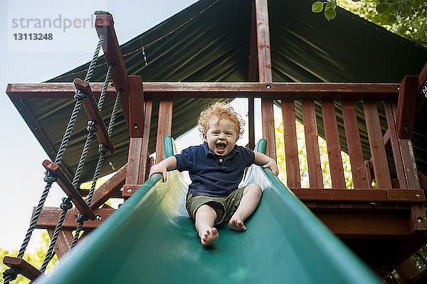 Niedrigwinkel-Porträt eines glücklichen kleinen Jungen  der schreit  während er auf dem Hof rutscht