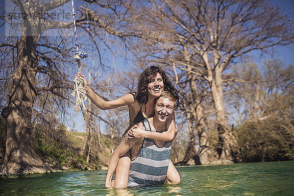 Porträt einer fröhlichen Freundin  die an einem sonnigen Tag ihrem Freund im Fluss vor kahlen Bäumen Huckepack gibt