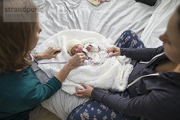 Hochwinkelaufnahme einer Hebamme  die ein neugeborenes Mädchen untersucht  während die Mutter zu Hause im Bett sitzt