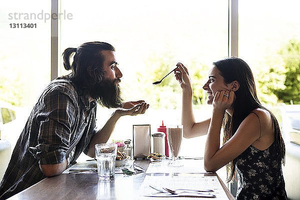 Seitenansicht einer glücklichen Frau  die einem Mann einen Löffel zeigt  während sie im Café sitzt