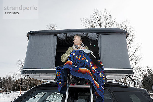 Frau strickt  während sie im Winter im Dachzelt auf dem Auto sitzt