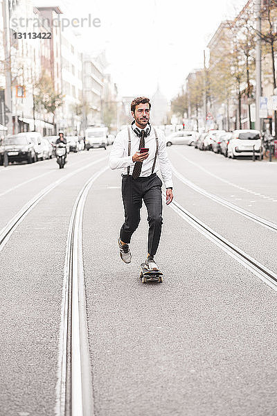 Geschäftsmann hält Telefon  während er auf einer Straße in der Stadt Skateboard fährt