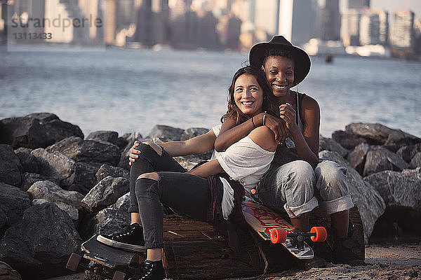 Freundinnen umarmen sich  während sie mit Skateboards am felsigen Ufer sitzen