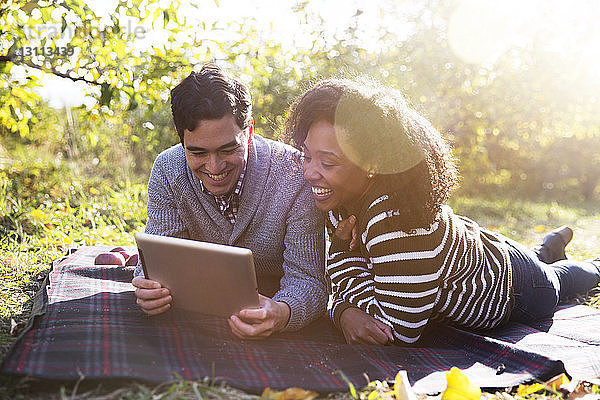 Paar benutzt einen Tablet-Computer  während es sich auf einer Decke im Obstgarten entspannt