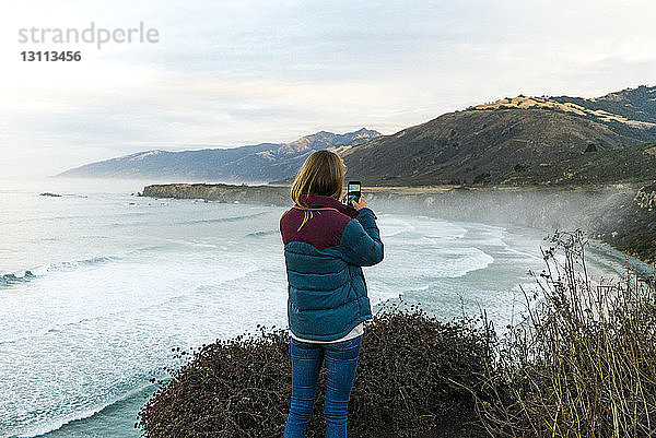 Rückansicht einer Frau  die einen Berg fotografiert  während sie auf einem Hügel gegen das Meer steht