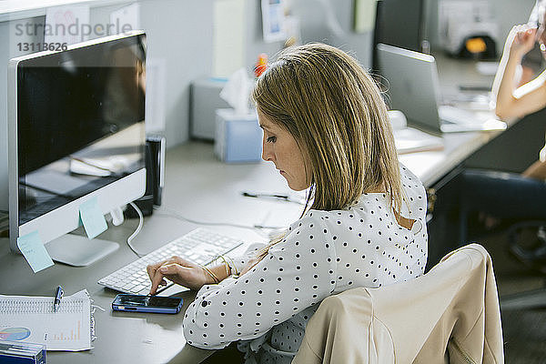 Geschäftsfrau benutzt Mobiltelefon  während sie mit einem Kollegen am Schreibtisch sitzt