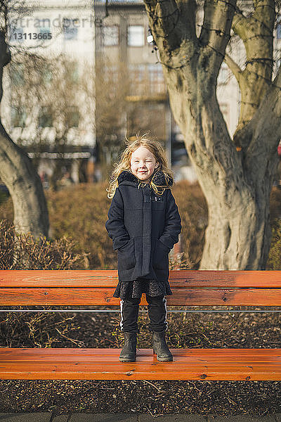 Ganzfigur eines Mädchens in voller Länge mit zwinkernden Händen in den Manteltaschen  das auf einer Parkbank steht