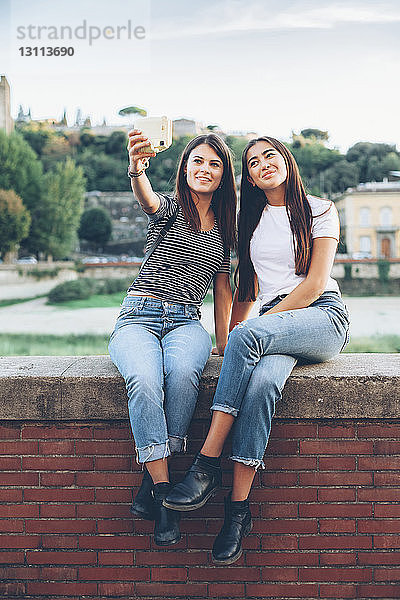 Glückliche Freundinnen in voller Länge  die sich mit einer Sofortbildkamera selbst fotografieren  während sie auf einer Stützmauer im Park sitzen