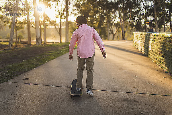 Rückansicht eines Jungen  der bei Sonnenuntergang auf einem Fußweg im Park Skateboard fährt