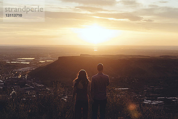 Rückansicht eines jungen Paares  das bei Sonnenuntergang auf dem Feld gegen den Himmel steht