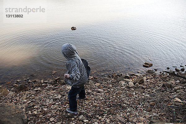 Seitenansicht eines Jungen mit Kapuzenhemd  der einen Stein in den See wirft