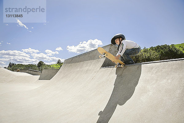 Niedrigwinkel-Ansicht eines Jungen  der ein Skateboard auf der Rampe gegen den blauen Himmel hält
