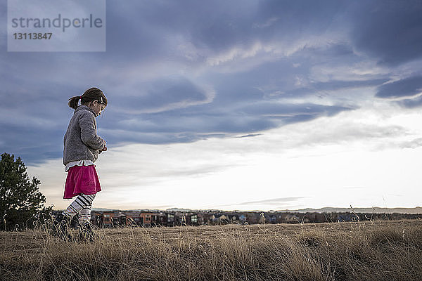 Niedrigwinkelaufnahme eines Mädchens in warmer Kleidung  das bei Sonnenuntergang auf einem Grasfeld vor bewölktem Himmel geht