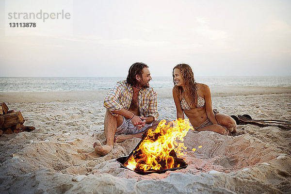 Glückliches Paar unterhält sich am Strand am Lagerfeuer im Sand sitzend