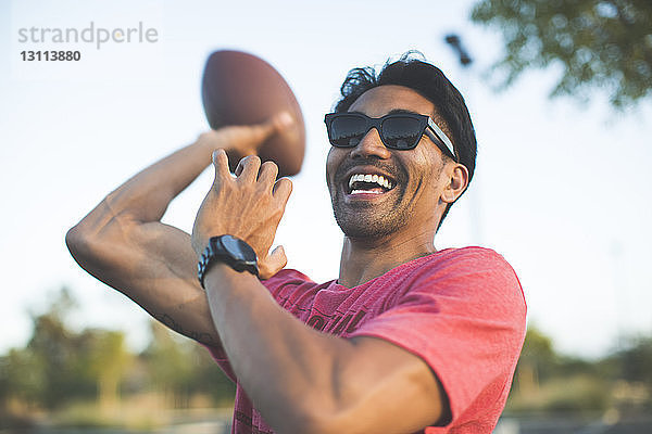 Fröhlicher Mann mit Sonnenbrille spielt mit American Football gegen den Himmel