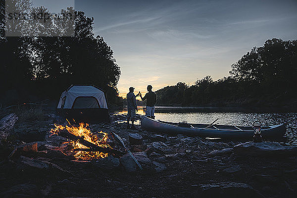 Rückansicht von männlichen Freunden  die während des Sonnenuntergangs auf dem Campingplatz am See auf Getränke anstoßen