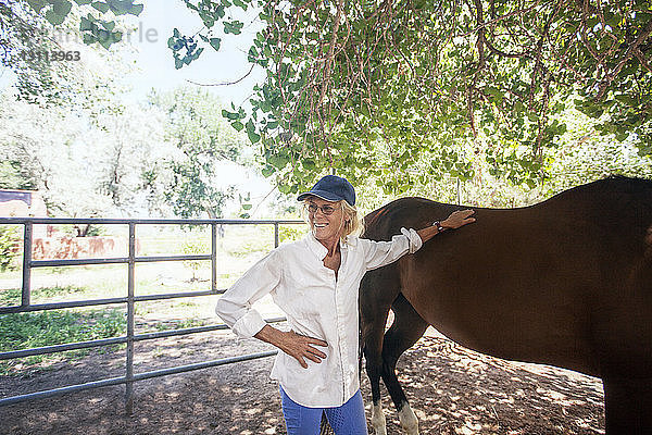 Zuversichtliche ältere Frau steht mit Pferd an Baum