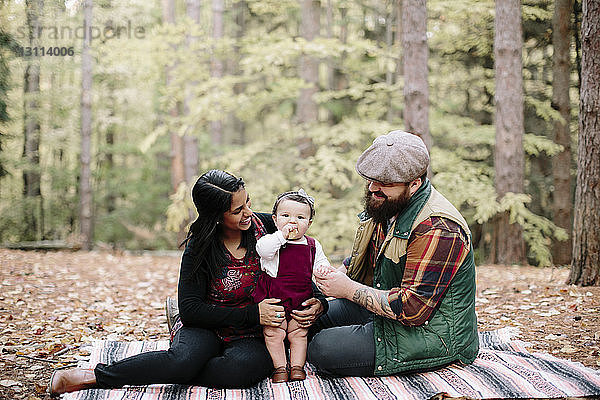 Glückliche Eltern schauen der Tochter beim Essen zu  während sie sich auf einer Decke im Wald entspannen