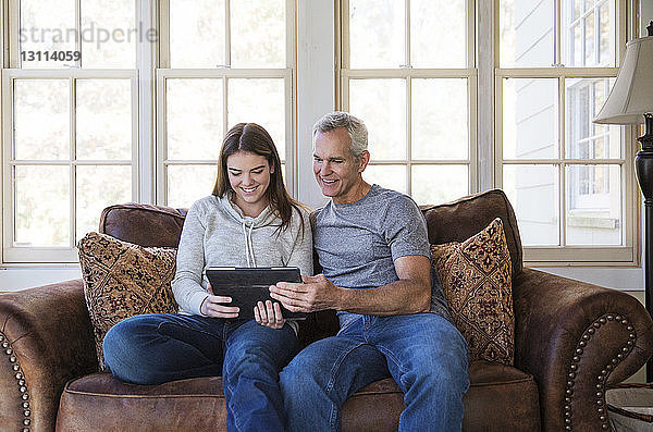 Glückliche Teenagerin betrachtet Tablette  während sie zu Hause mit Großvater auf dem Sofa sitzt
