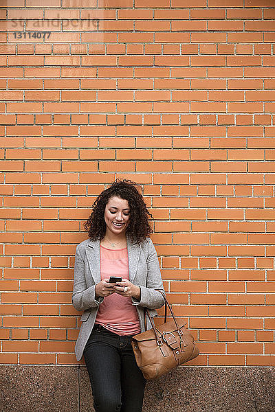 Glückliche Geschäftsfrau benutzt Smartphone  während sie an einer Ziegelmauer steht