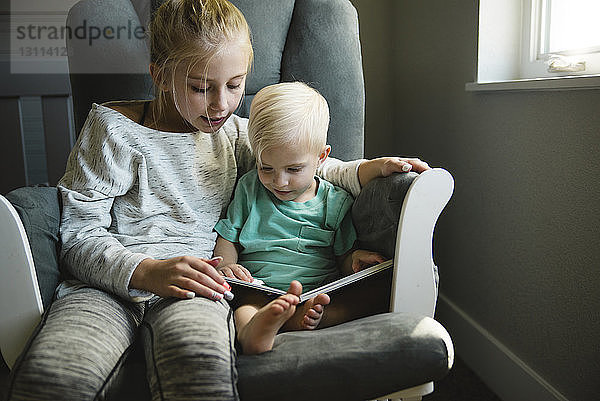 Schwester und Bruder schauen sich ein Buch an  während sie zu Hause auf einem Stuhl sitzen