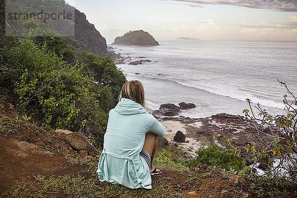 Rückansicht einer Frau  die auf einem Hügel sitzend auf das Meer blickt