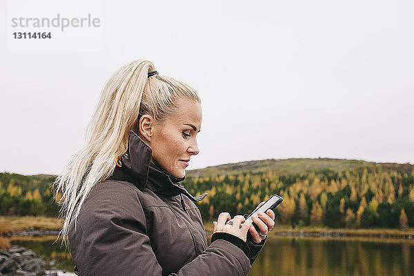 Seitenansicht einer Frau  die ein Smartphone am See gegen den klaren Himmel im Herbst benutzt