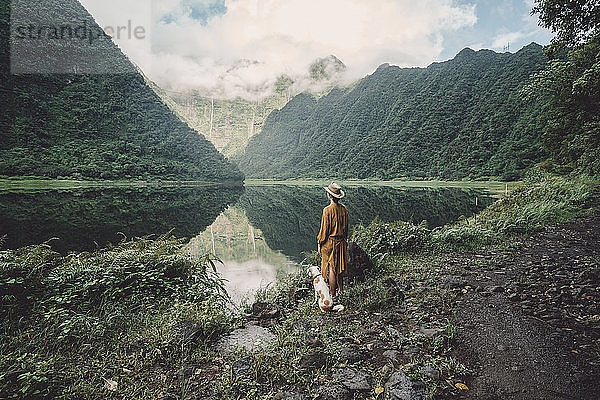 Rückansicht einer am Seeufer stehenden Frau mit Hund vor bewölktem Himmel im Wald