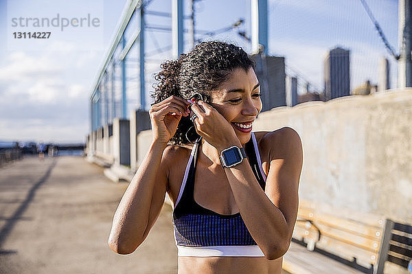 Lächelnde Athletin beim Einstellen des Headsets beim Gehen auf dem Fußweg gegen den Himmel