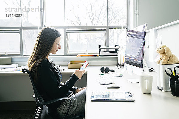 Geschäftsfrau benutzt Smartphone  während sie im Büro auf einem Stuhl am Schreibtisch sitzt