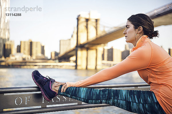 Nachdenkliche Athletin beim Beinstrecken auf der Promenade mit der Brooklyn Bridge im Hintergrund