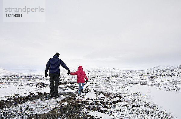 Rückansicht eines Vaters und einer Tochter  die sich an den Händen halten  während sie auf einer verschneiten Landschaft gegen den Himmel gehen