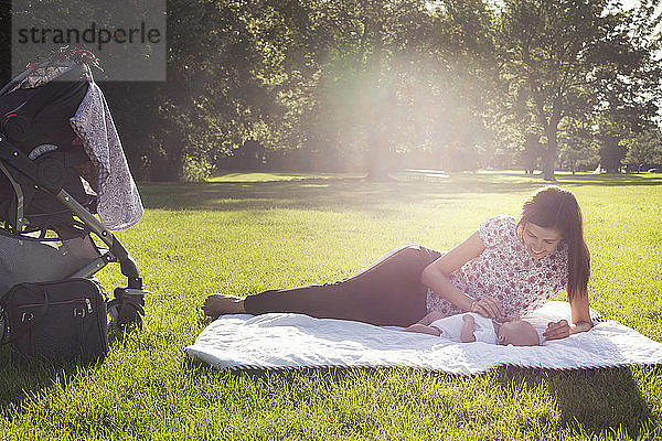 Glückliche Mutter spielt mit dem Baby  während sie sich an einem sonnigen Tag auf einem Grasfeld ausruht