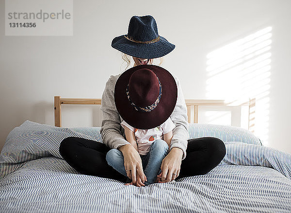 Glückliche Mutter mit Tochter  die zu Hause auf dem Bett sitzt und Hüte trägt