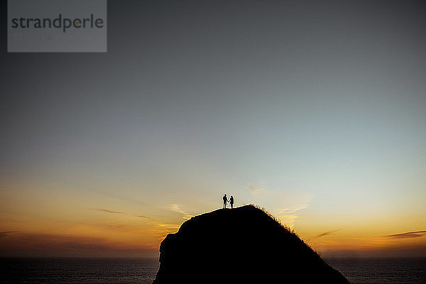 Fernansicht eines Paares  das bei Sonnenuntergang auf einer Silhouettenklippe gegen den Himmel am Meer steht