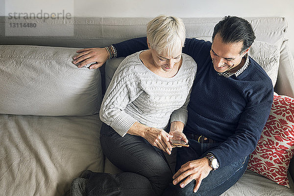 Schrägansicht eines älteren Paares  das zu Hause auf dem Sofa sitzend auf ein Smartphone schaut