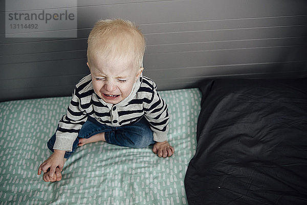 Hochwinkelaufnahme eines weinenden Jungen  der zu Hause auf dem Bett sitzt