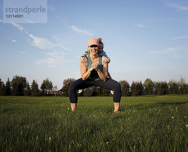 Glückliche Mutter trägt ihre Tochter  während sie auf einem Grasfeld im Park gegen den Himmel trainiert