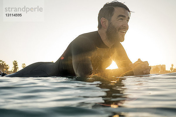 Glücklicher männlicher Surfer  der bei Sonnenuntergang auf einem Surfbrett im Meer liegt