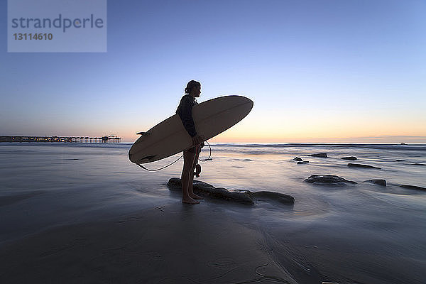 Frau hält Surfbrett  während sie am Ufer vor klarem Himmel steht