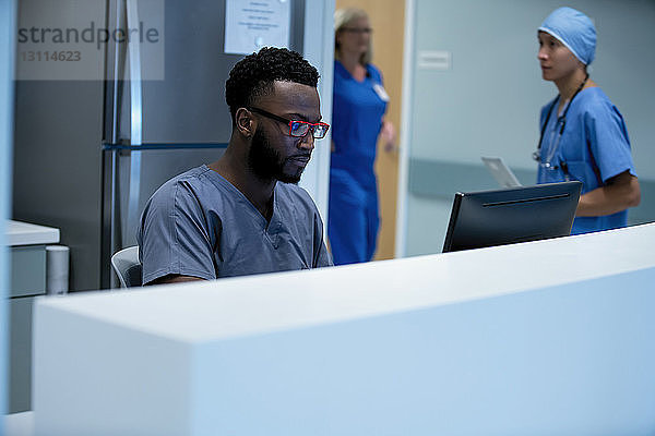 Zuversichtlicher Arzt am Desktop-Computer am Krankenhausempfang mit Kollegen  die im Hintergrund arbeiten