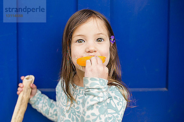 Porträt eines süßen Mädchens  das orange vor blauer Tür isst