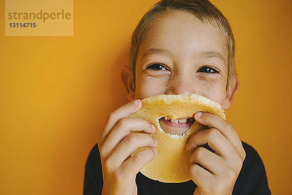 Glücklicher Junge schaut weg  während er zu Hause Brot mit Loch gegen eine orangefarbene Wand hält