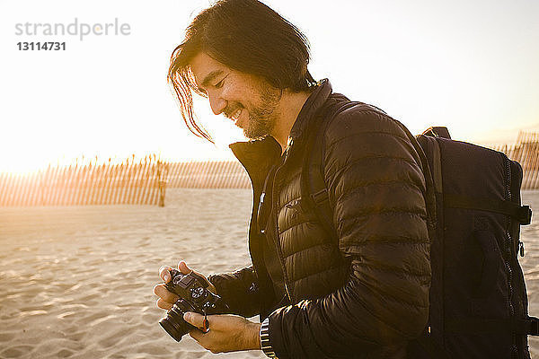 Seitenansicht eines am Strand stehenden lächelnden Mannes mit Rucksack und Kamera